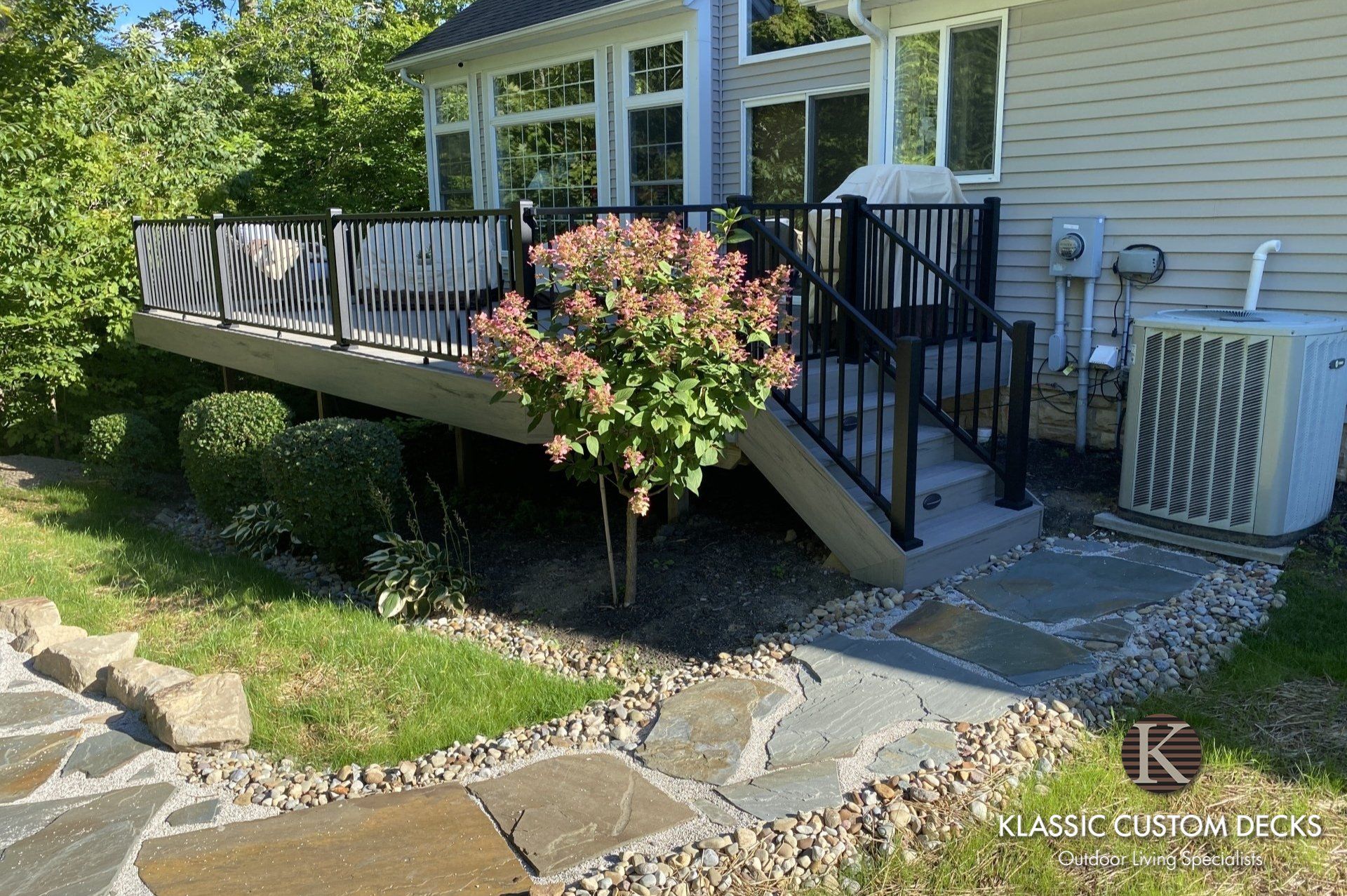 Back deck with stairs, black railings, pink flowering shrubs, and stone walkway beside a house
