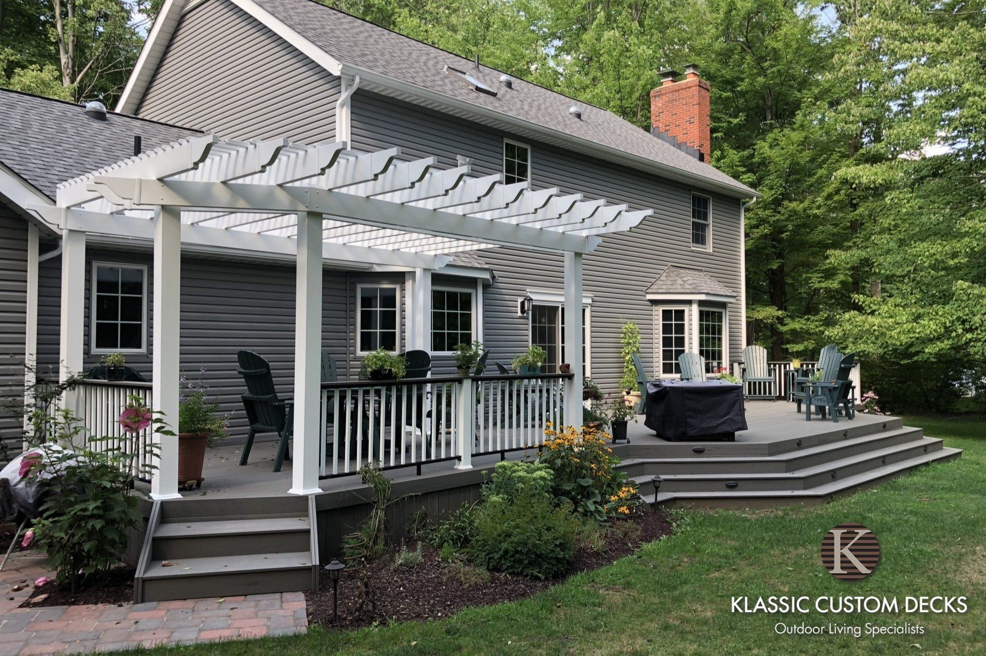 White house with gray roof, covered front porch, steps, patio seating, and landscaped garden in summer.
