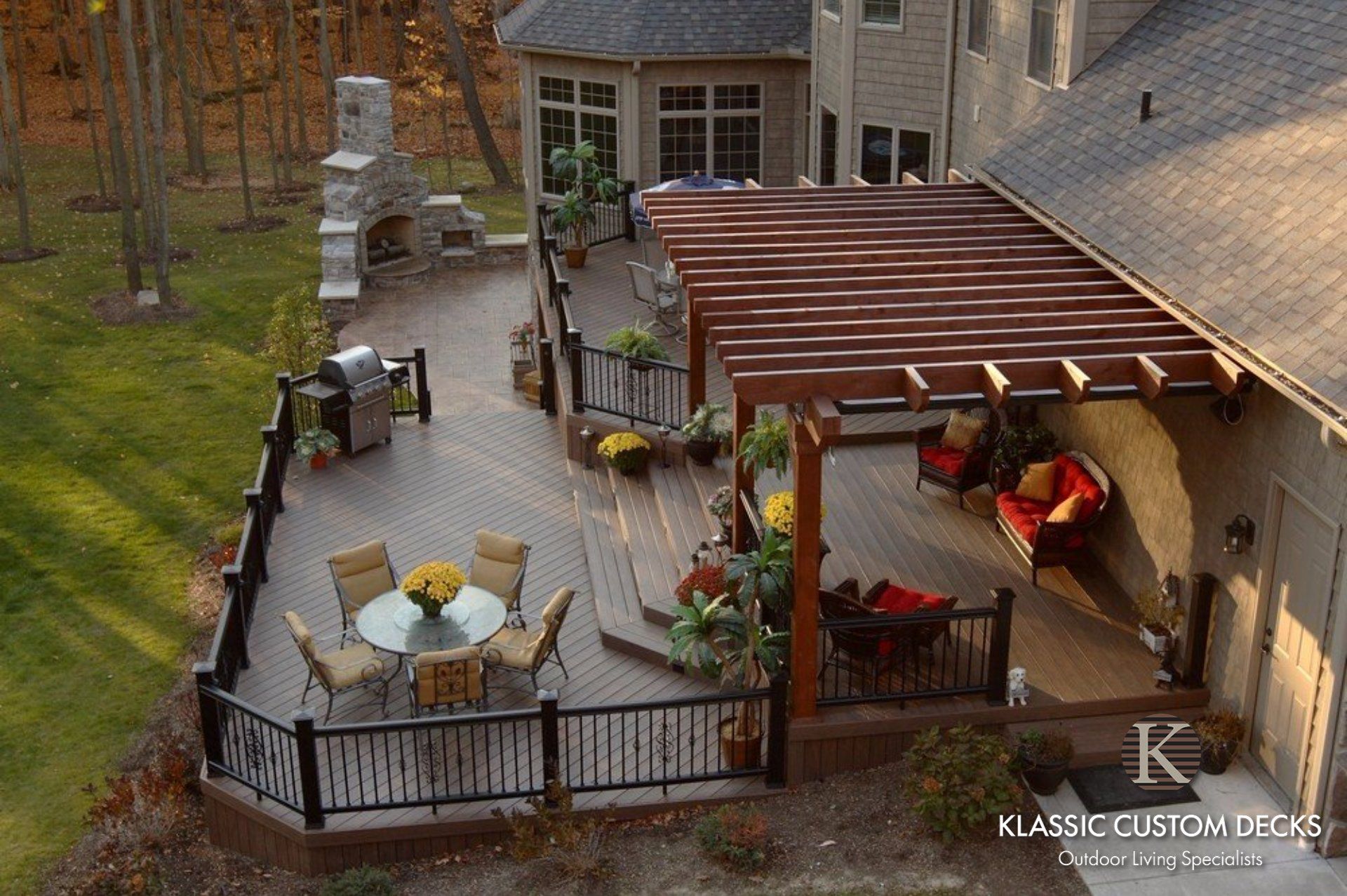 Wood deck patio with black railing, round table, and brown pergola beside a stone house and woods