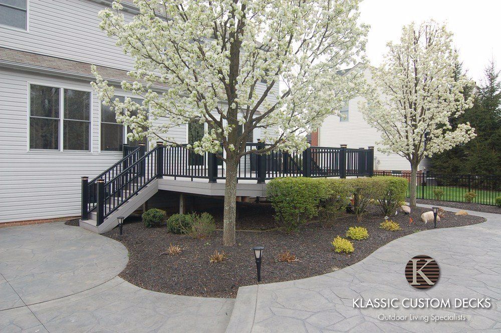 Front porch with black railing, flowering trees, and curved stone landscaping beside a white house