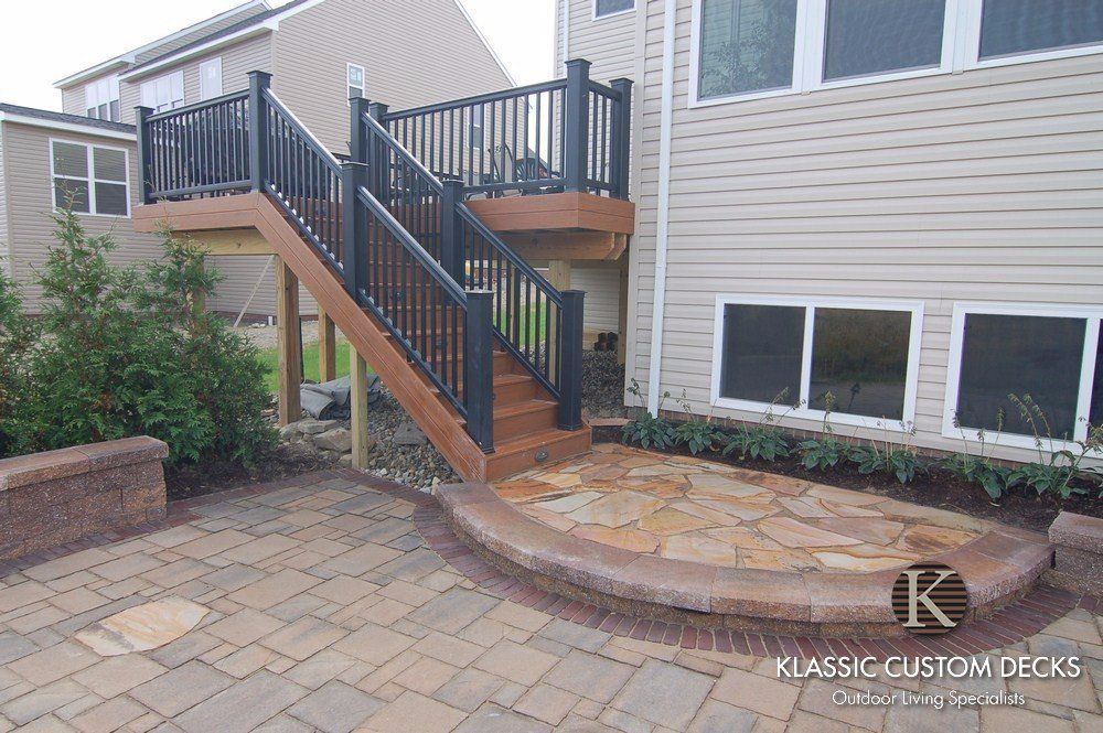 Backyard deck with stairs, black railings, and brick paver patio beside a house