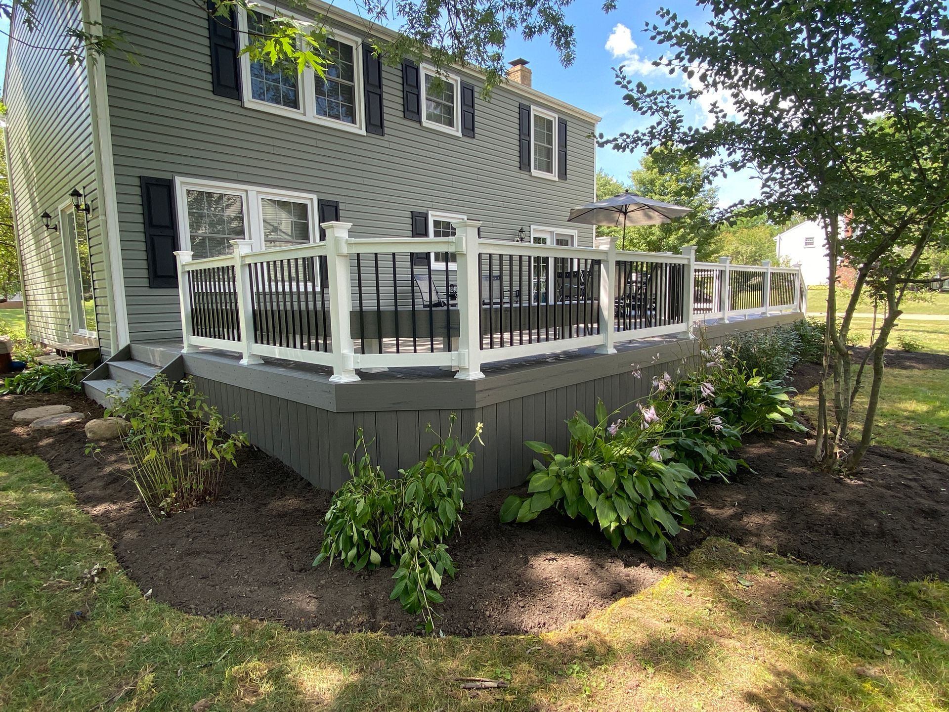 Gray house with white-railed deck and steps, surrounded by landscaped shrubs and trees