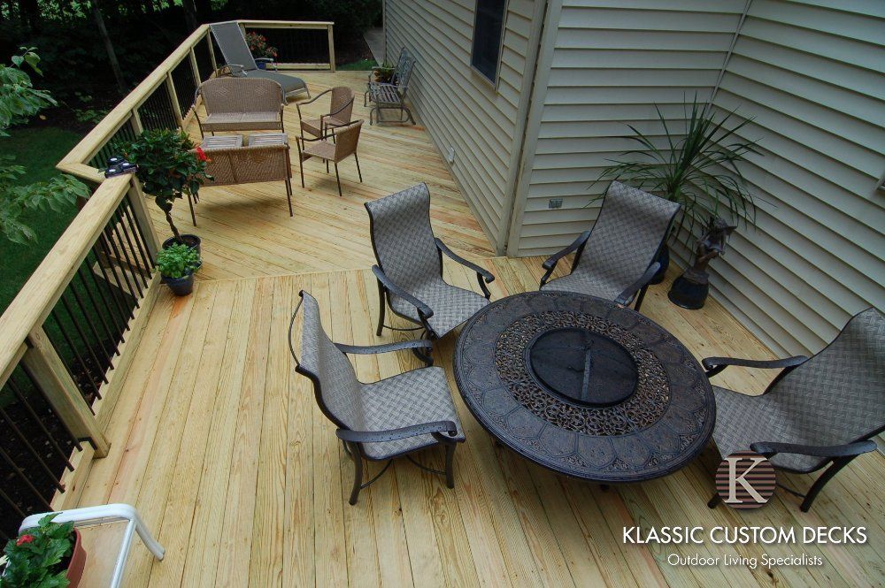 Wooden backyard deck with round fire pit table, wicker chairs, and potted plants along the house siding
