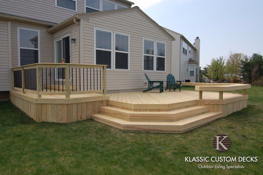 Wood deck with steps and railing beside a beige house, featuring a patio area with chairs.