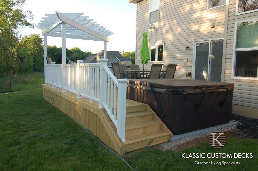 White pergola and railing on a raised wood deck beside a house, with a hot tub and patio chairs