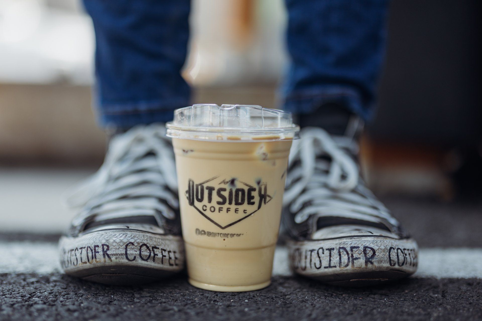 Iced coffee cup between two sneakered feet on pavement, with “The Outsider” branding.