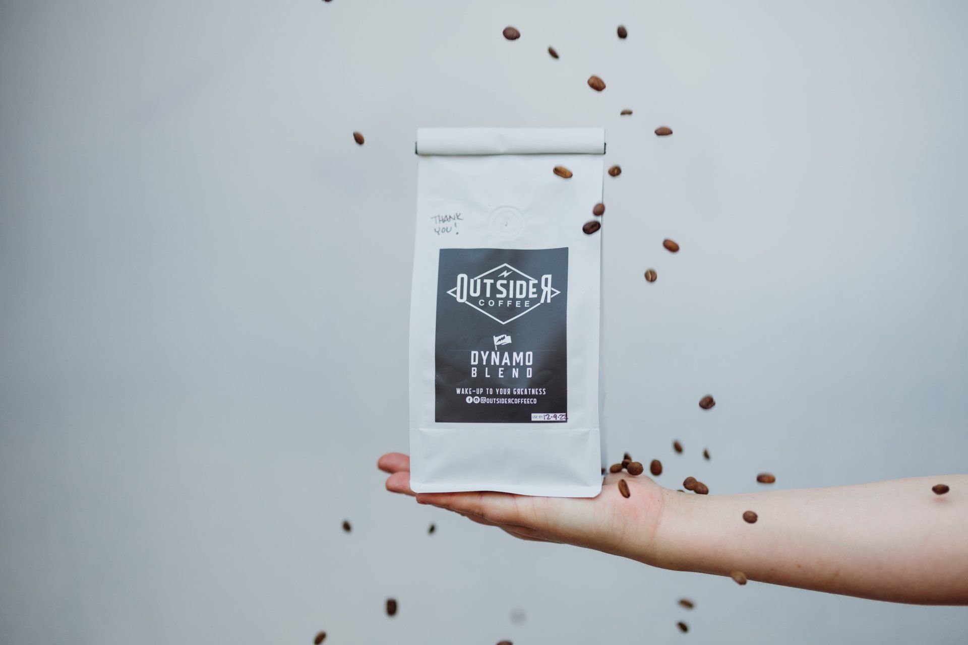 Hand holding a white coffee bag with beans floating around it on a light gray background