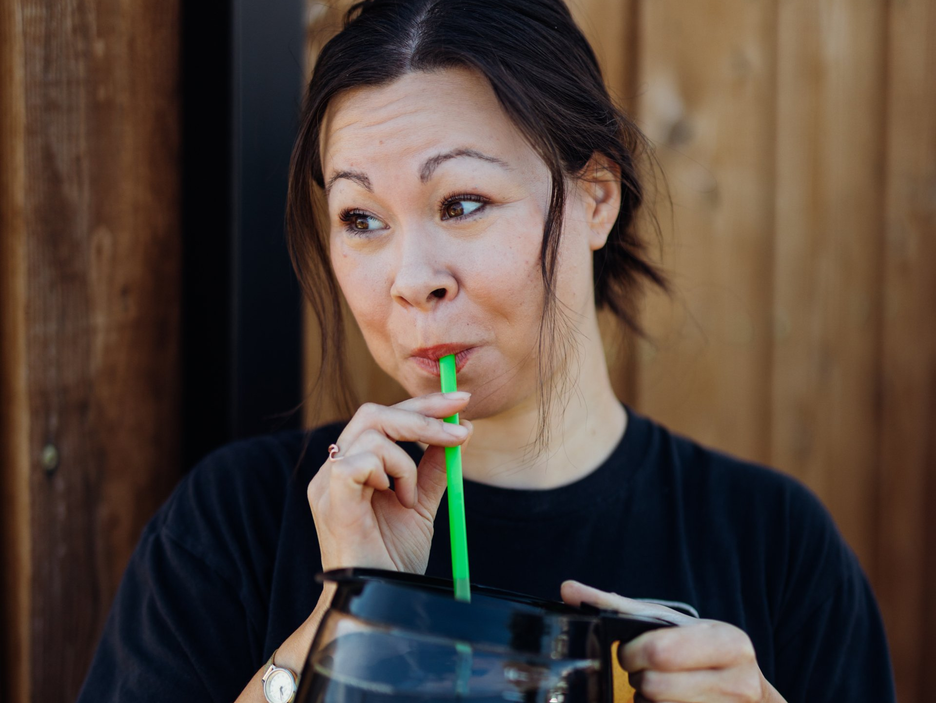 Woman in black shirt sipping a drink through a green straw, looking to the side with a playful expression.