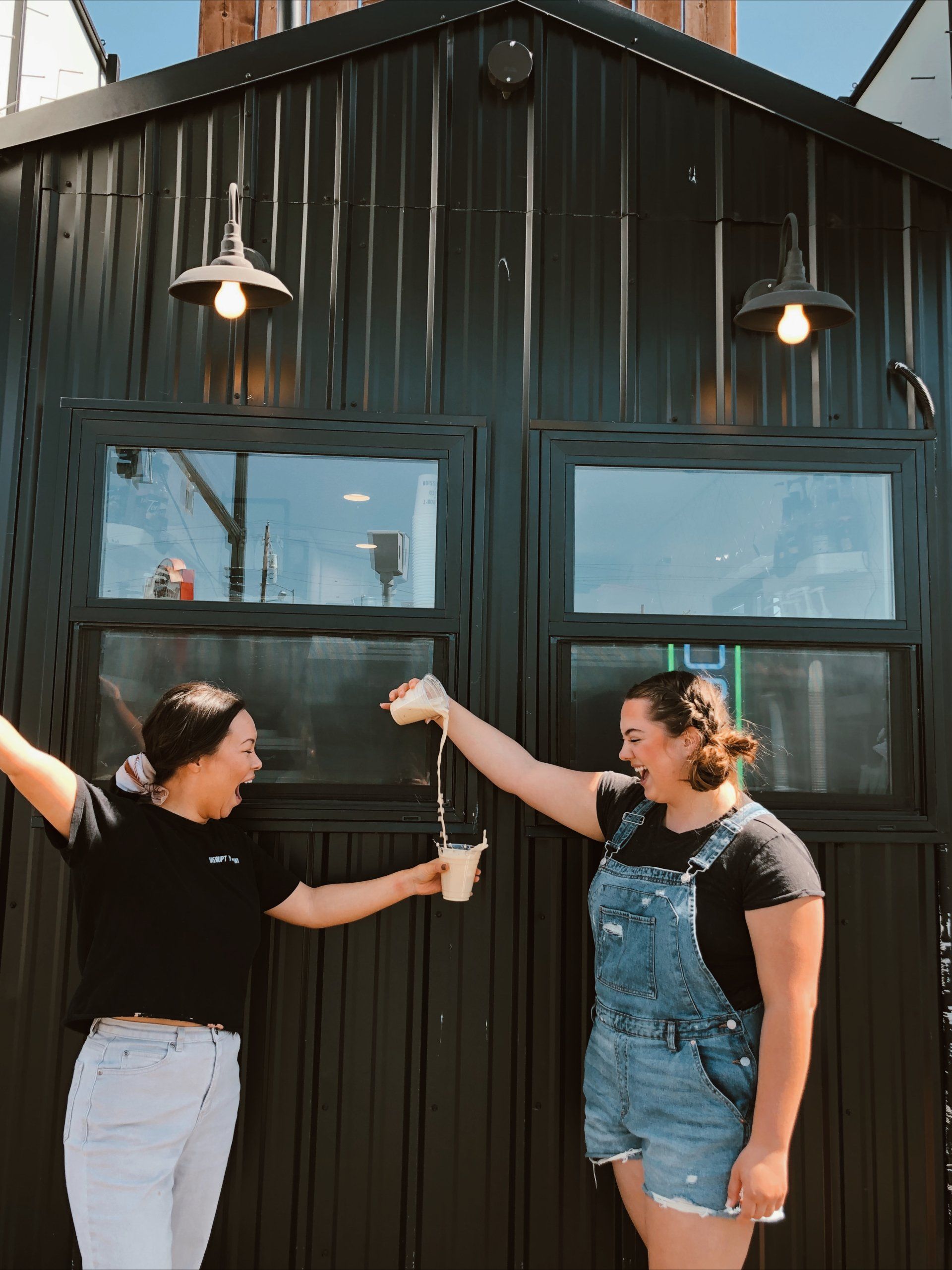 Two people standing by a dark metal building, smiling and pointing at each other under hanging lights