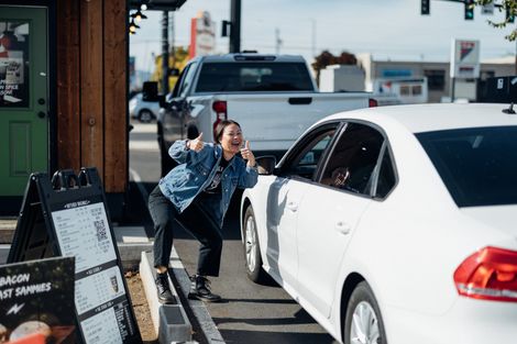 Woman crouches beside a white car at a curbside pickup window, talking to the driver.