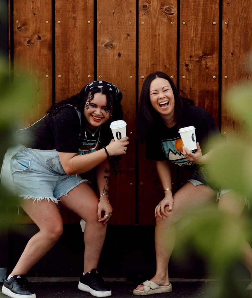 Two people crouch and smile, holding drinks in front of a wooden wall.