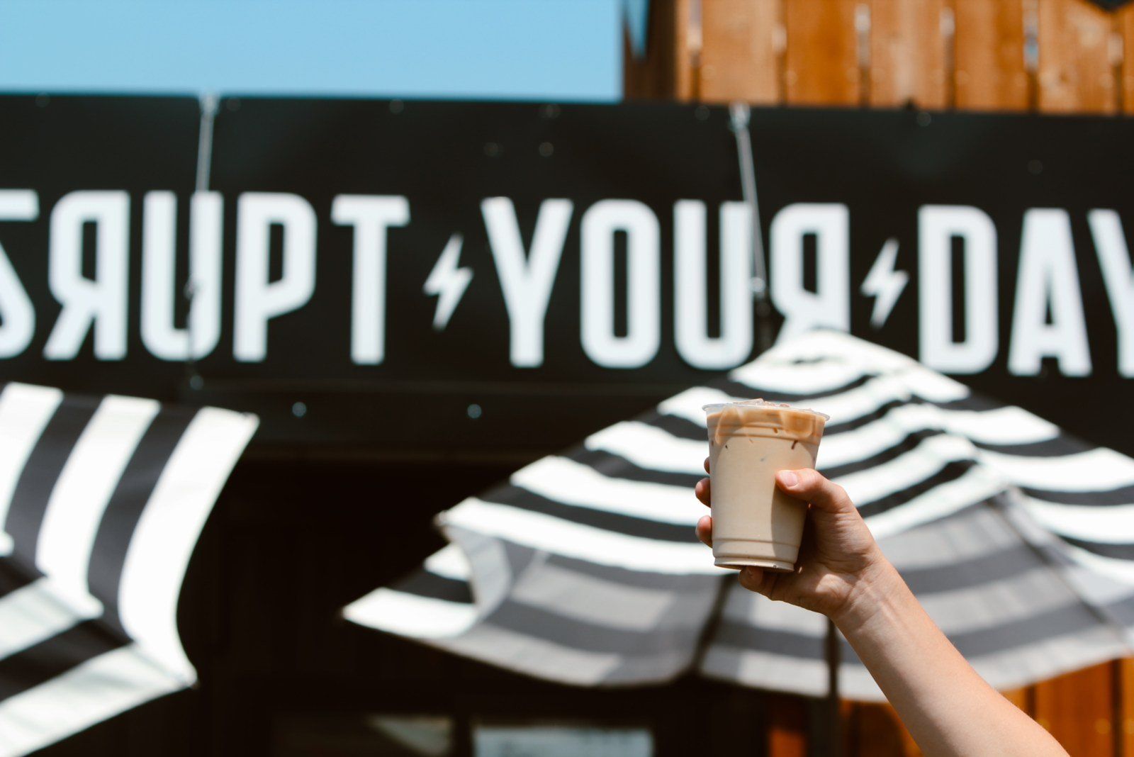 Hand holding a coffee cup in front of a black banner reading “YOUR DAY” with white striped awnings below