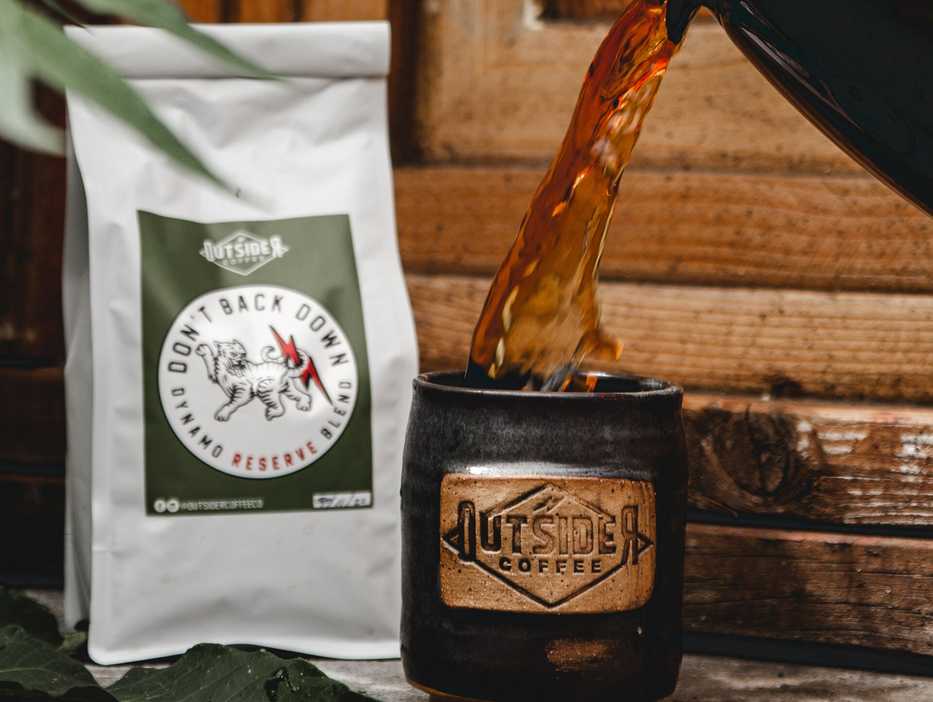 Coffee pouring into a black mug beside a white coffee bag on a rustic wooden table
