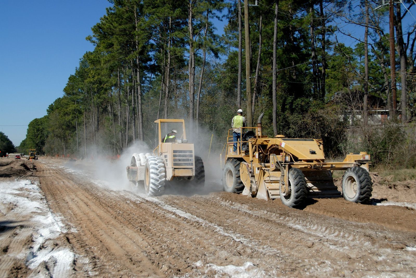 Two yellow road graders working on a dirt road, trees in the background, dust in the air.