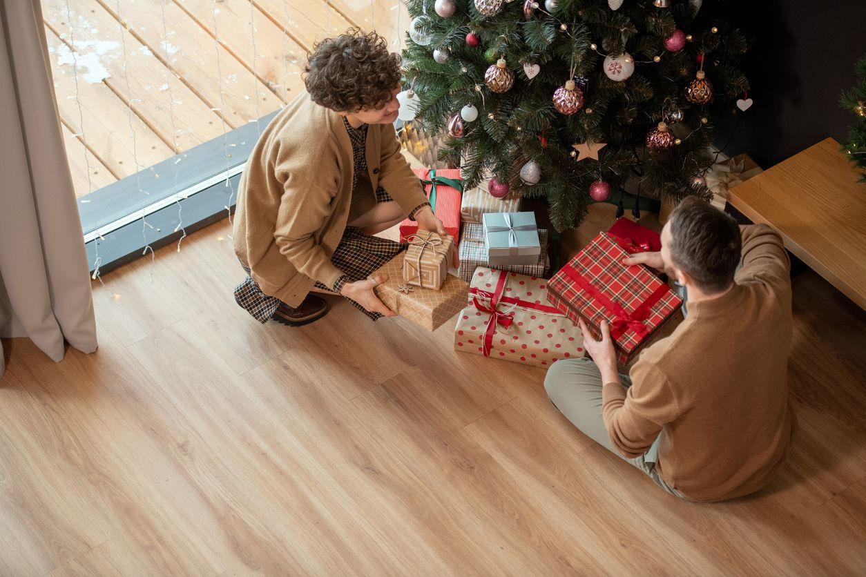 Two people sitting by a Christmas tree, wrapping gifts. Presents are under the tree.