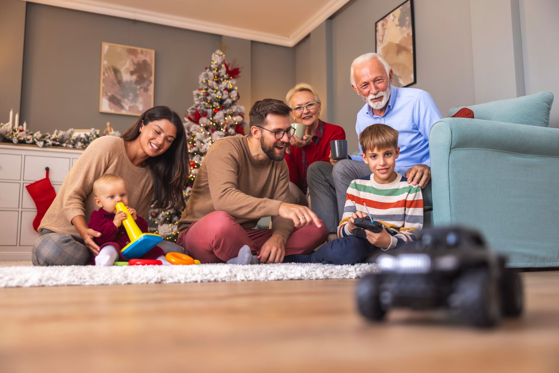 Family gathered at home; Christmas tree in background. Smiling people, baby with toy, boy playing game.