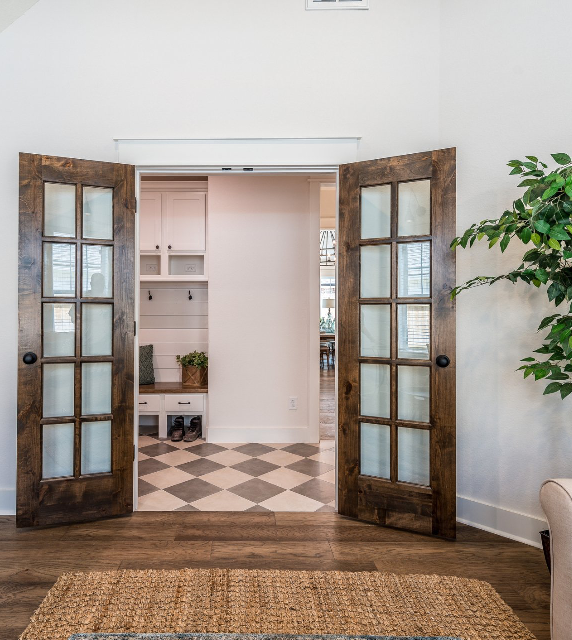 Open French doors leading to a bright room with checkered tile and shelving, with a potted plant on the right.