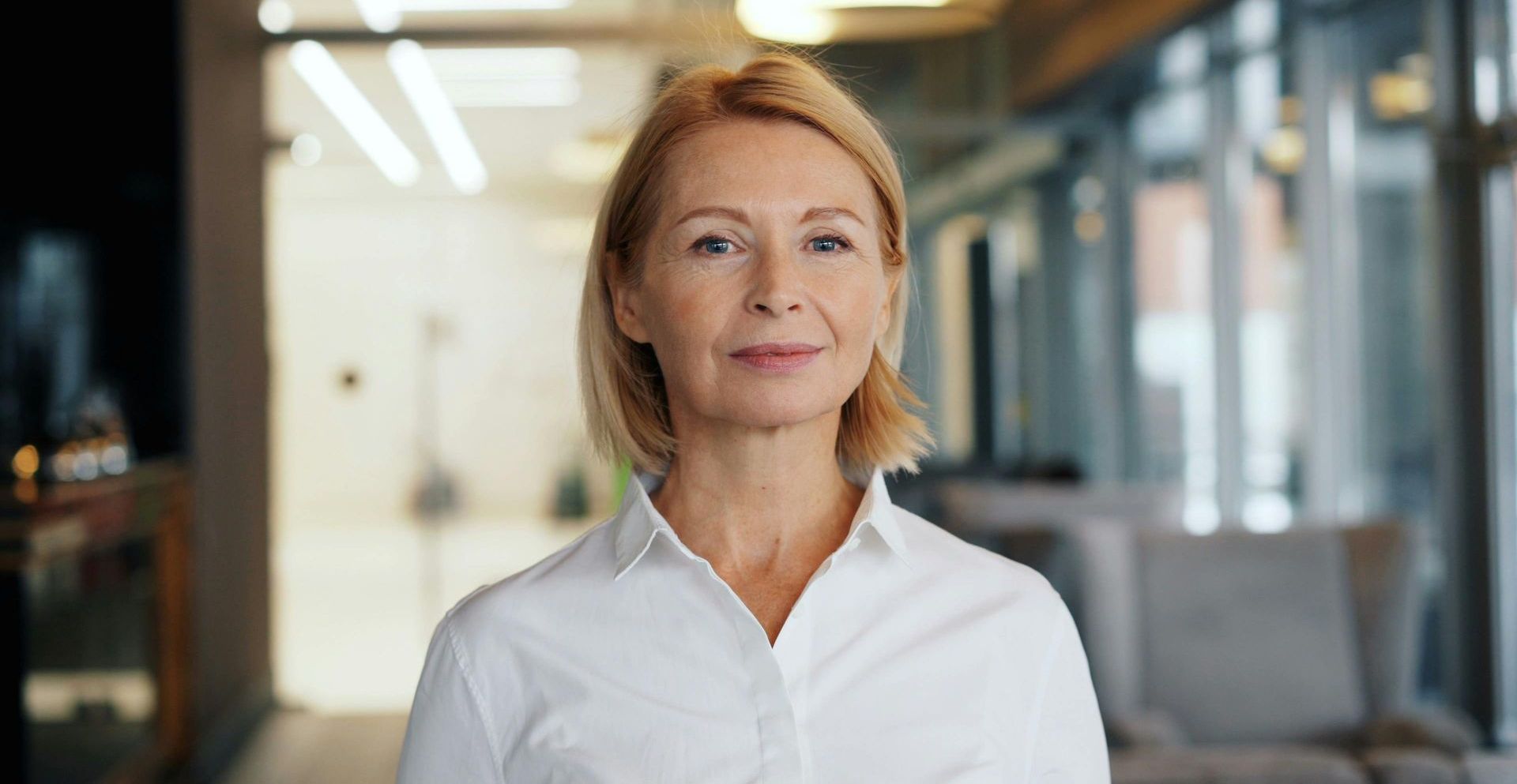 A woman in a white shirt is standing in a hallway and smiling at the camera.