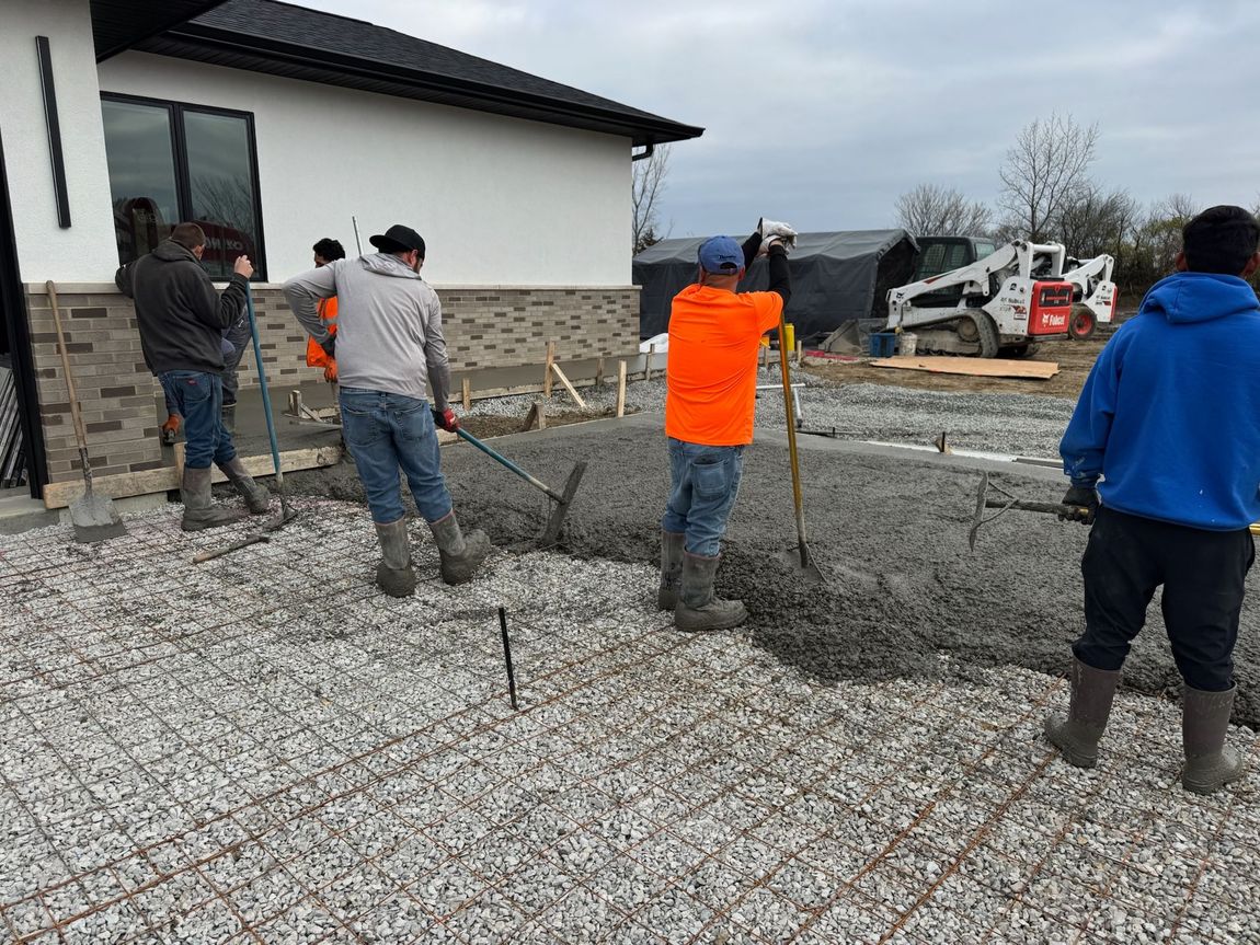 Construction workers leveling concrete on a prepared gravel base near a house.