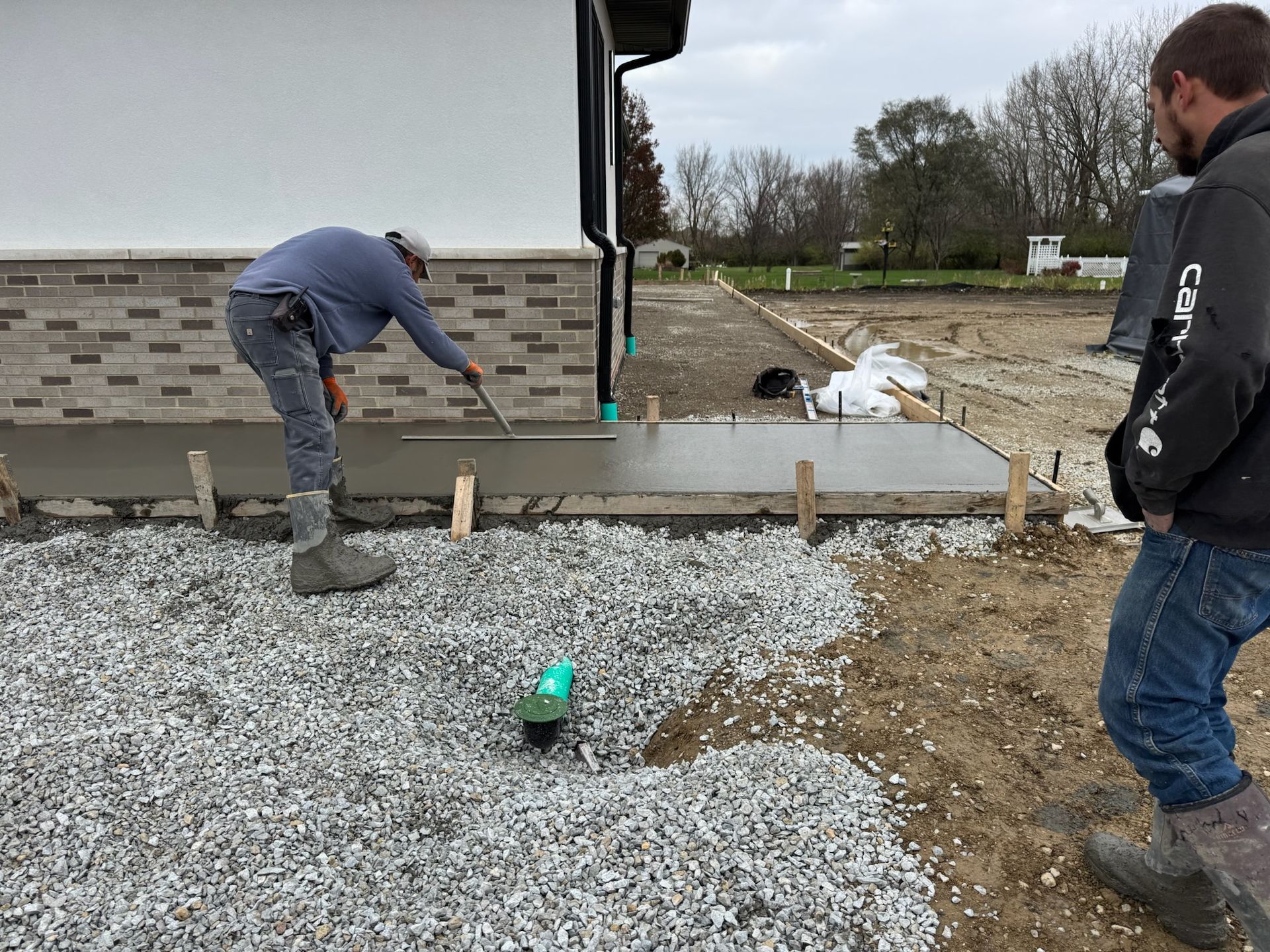 Two workers smoothing fresh concrete sidewalk. One uses a trowel. Gravel and dirt surround the work area. Two workers smoothing fresh concrete sidewalk. One uses a trowel. Gravel and dirt surround the work area.
