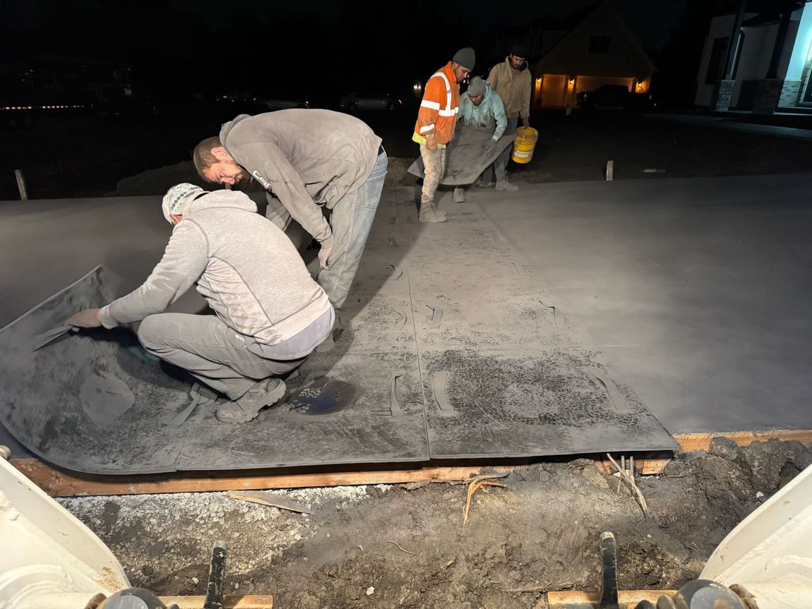 Workers smoothing concrete at night. One kneels, others carry a sheet and bucket in a dimly lit outdoor setting.