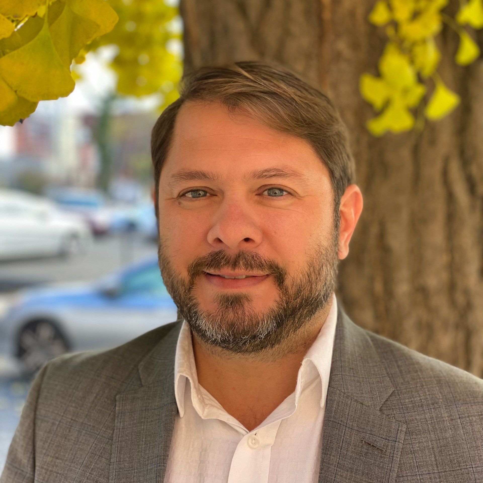 A man with a beard wearing a suit and white shirt is standing in front of a tree.