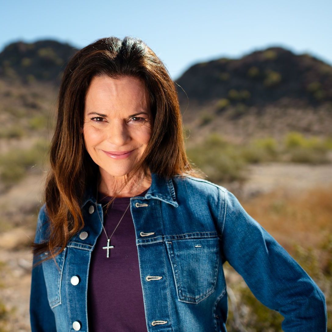 A woman wearing a denim jacket and a cross necklace is standing in the desert.