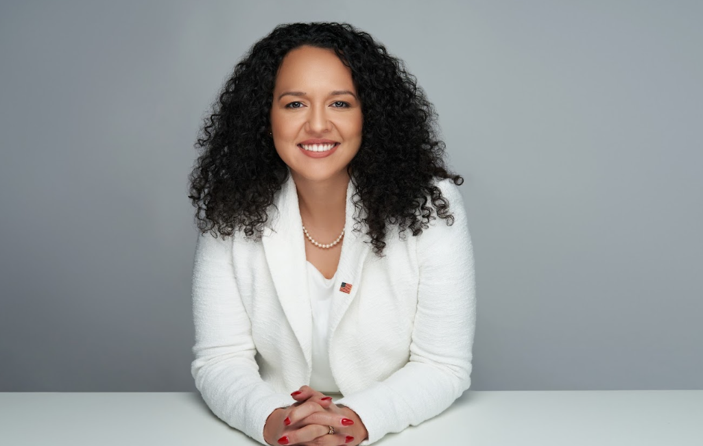 A woman is sitting at a table with her hands folded and smiling.