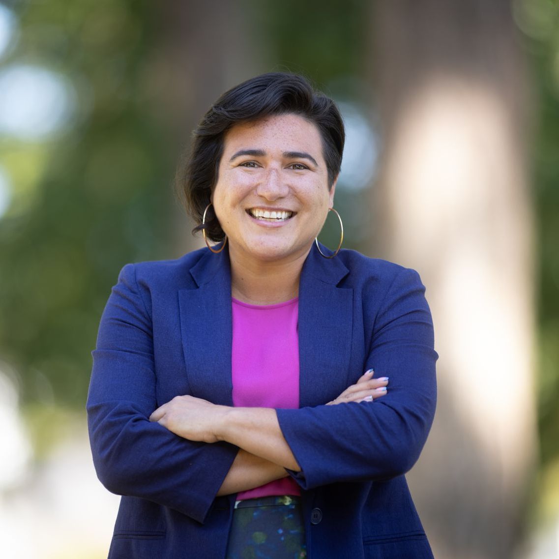 A woman in a blue jacket and pink shirt is smiling with her arms crossed.