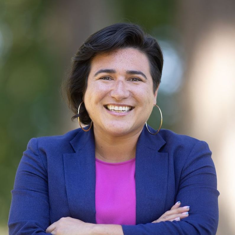 A woman in a blue jacket and pink shirt is smiling with her arms crossed.