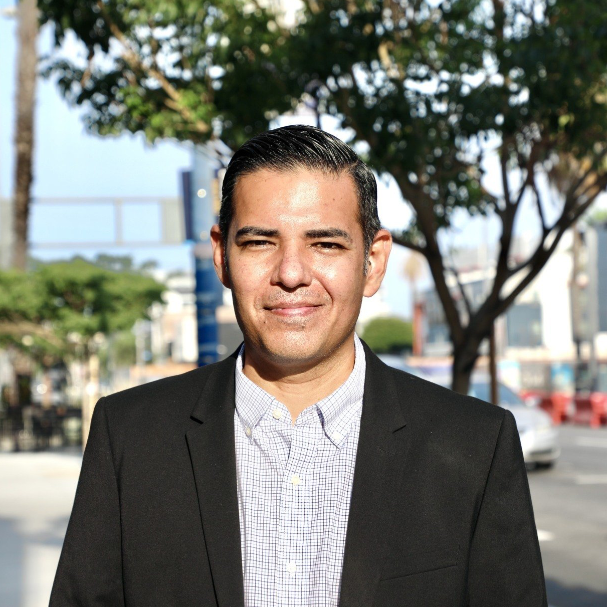 A man in a suit and shirt is standing in front of a tree.