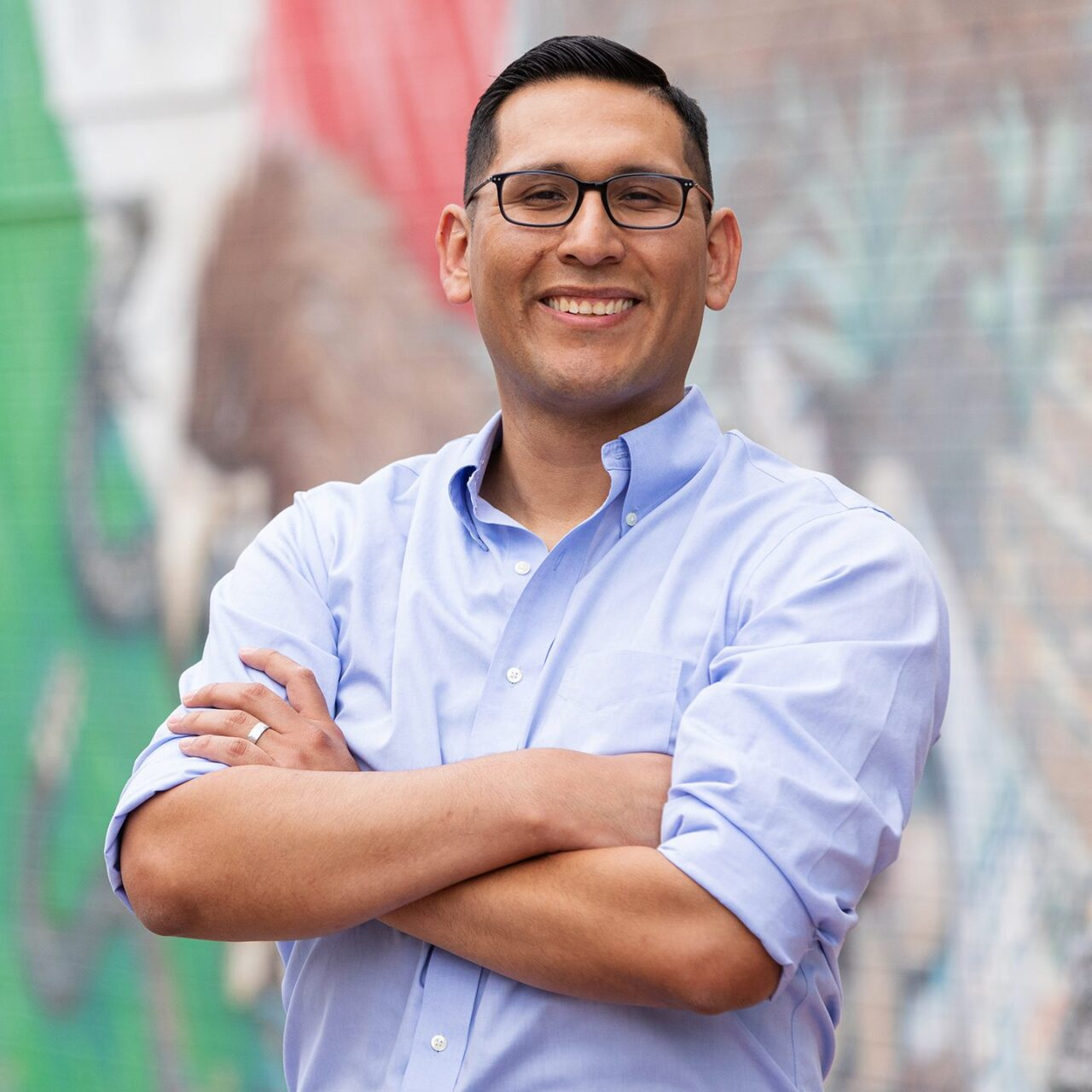 A man wearing glasses and a blue shirt is standing with his arms crossed and smiling.