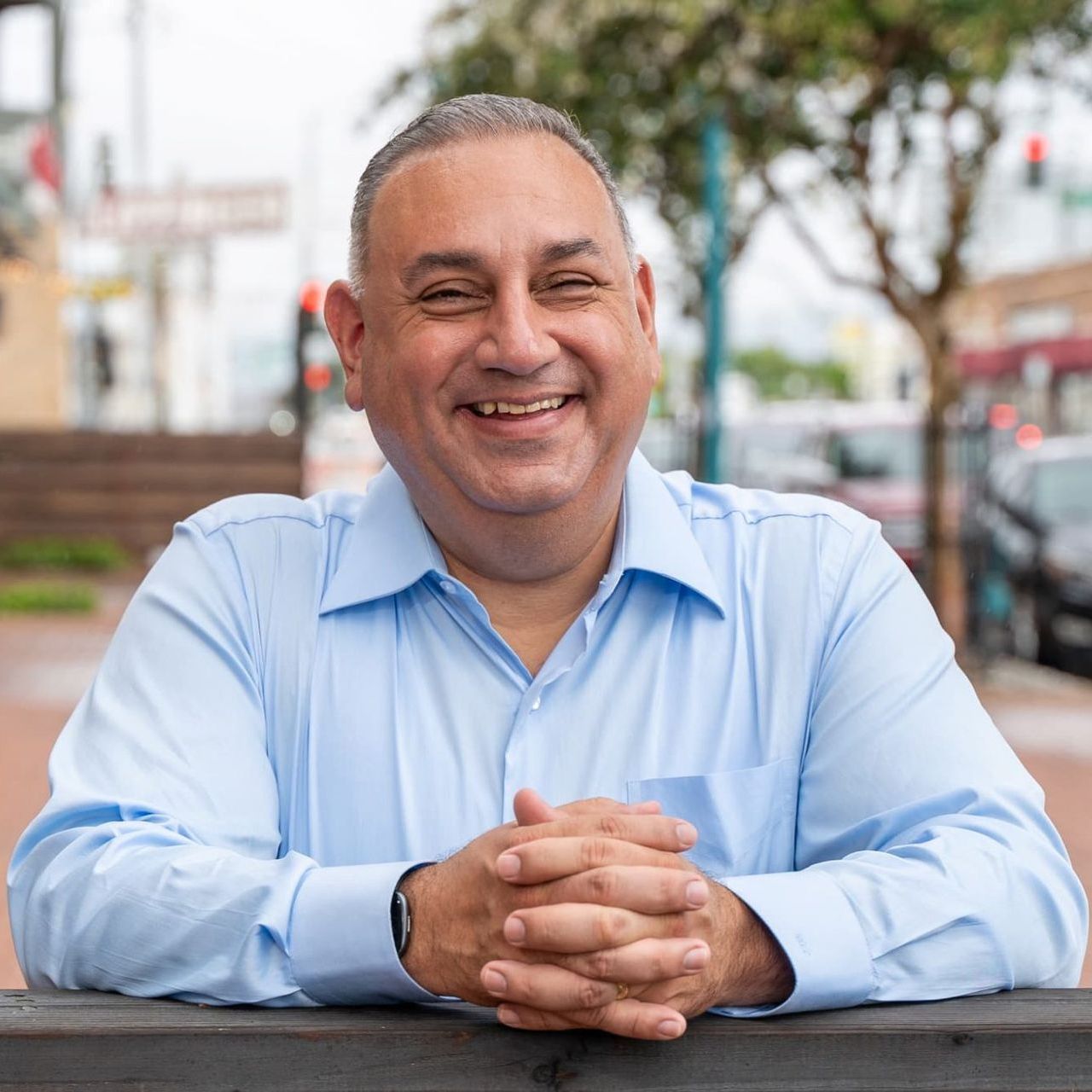 A man in a blue shirt is leaning on a railing with his hands folded and smiling.