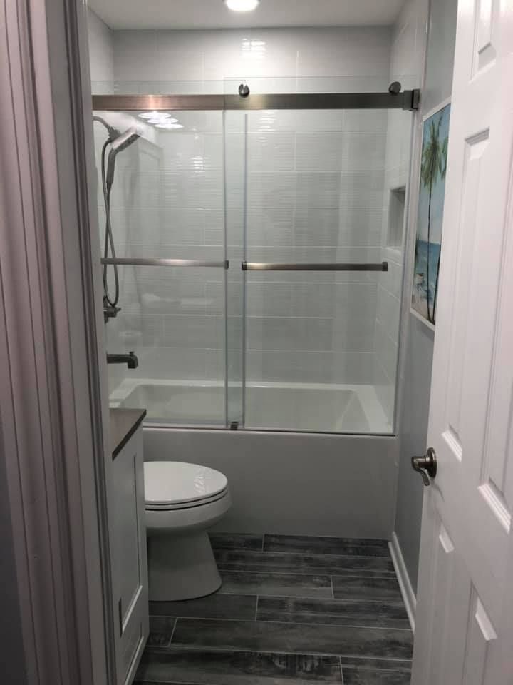 Small bathroom with shower, toilet, and gray wood-look floor. White tile walls, glass shower doors.