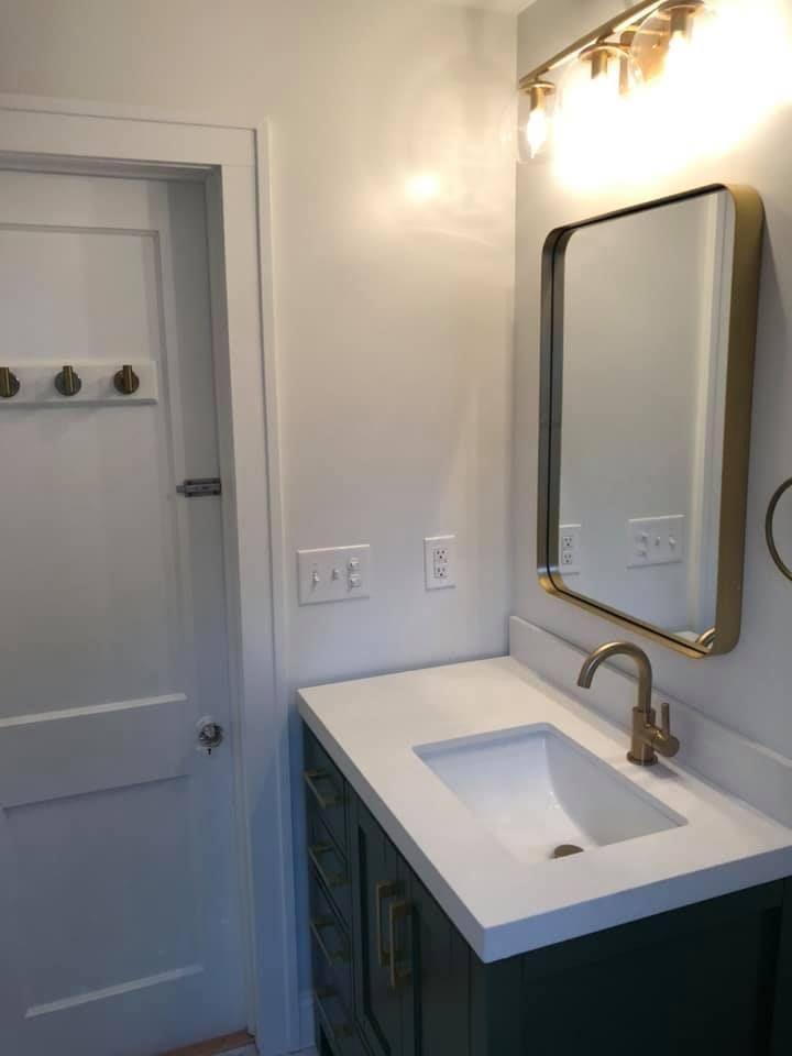 Bathroom with white countertop, green cabinet, gold faucet, mirror, and light fixture. White door with hooks.