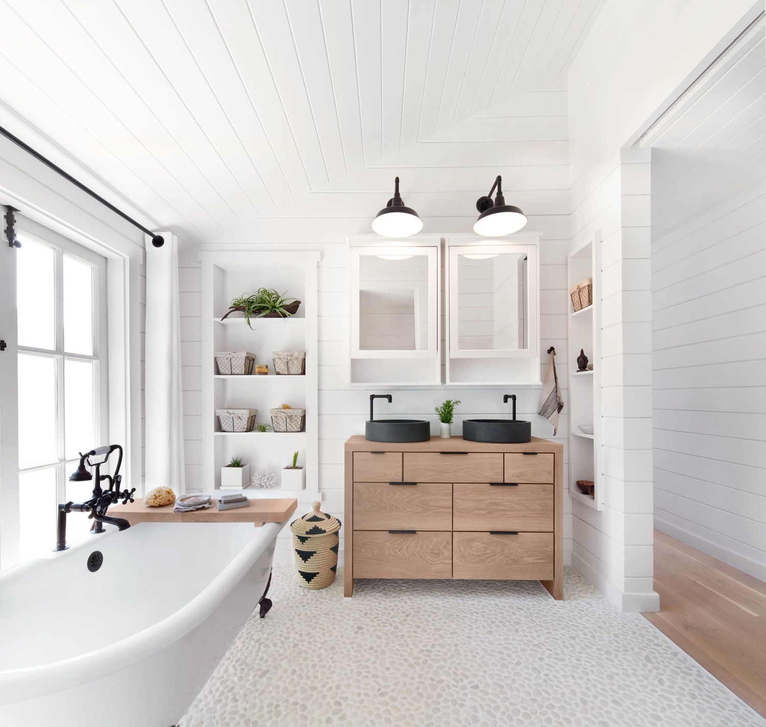 White bathroom with light wood vanity, black sinks, and soaking tub by window.