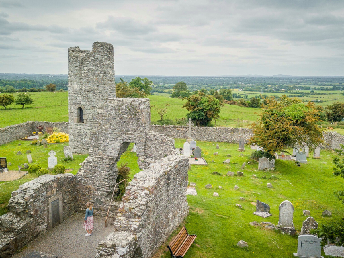 Stone ruins in a green field with a tall tower, scattered gravestones, and distant countryside views