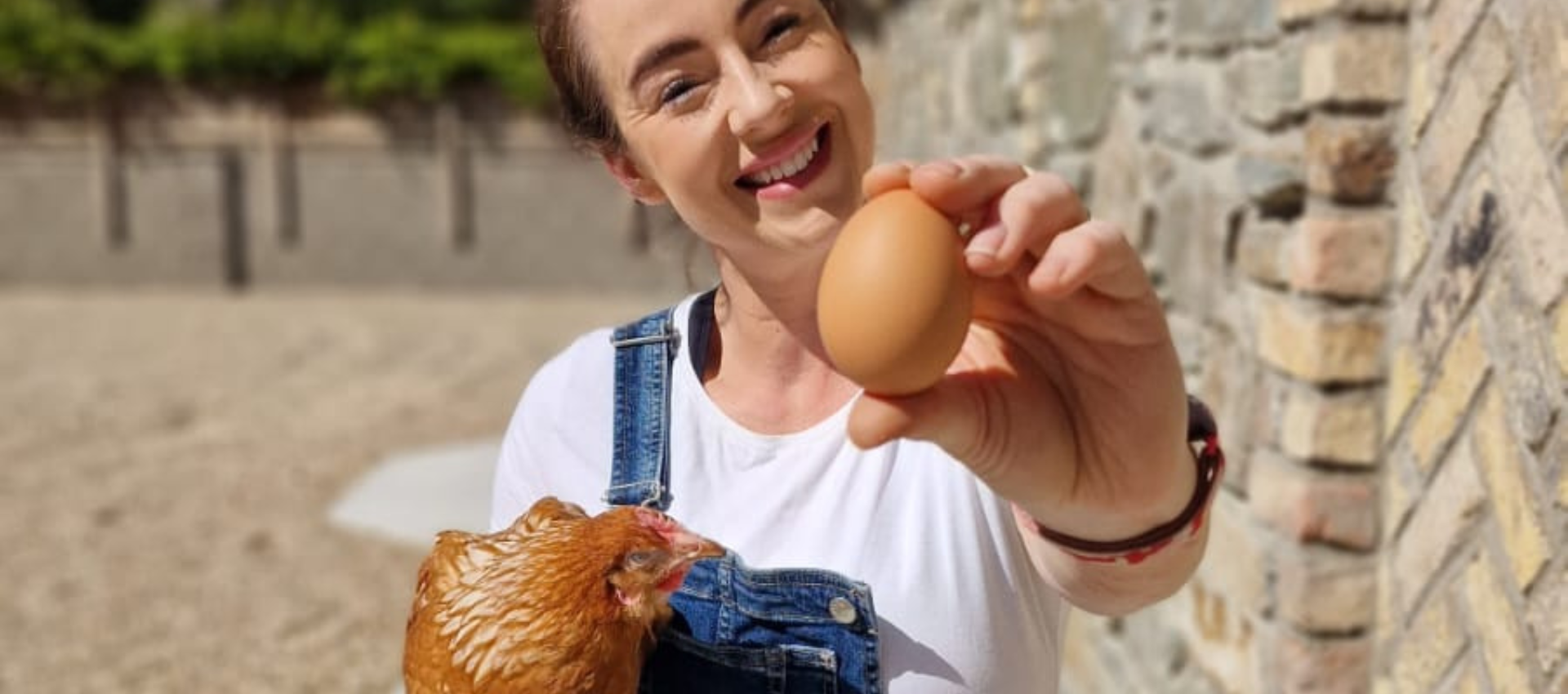 Woman holding up an egg and smiling outdoors with a croissant in the foreground