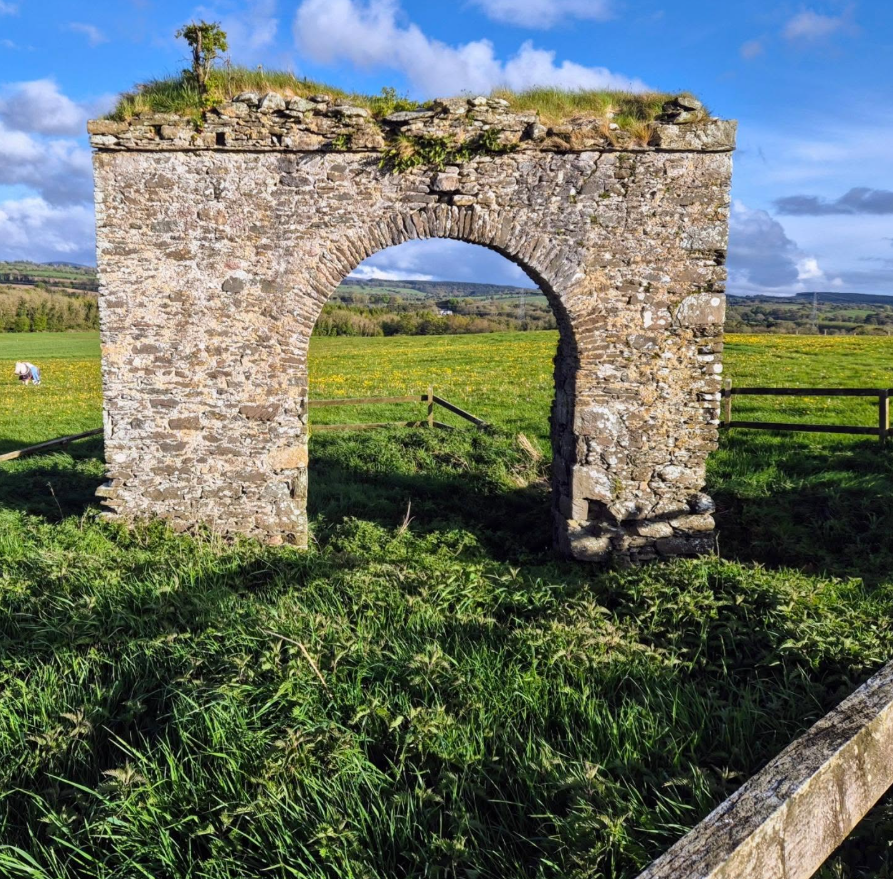 Stone ruin arch in a grassy field under a blue sky with clouds