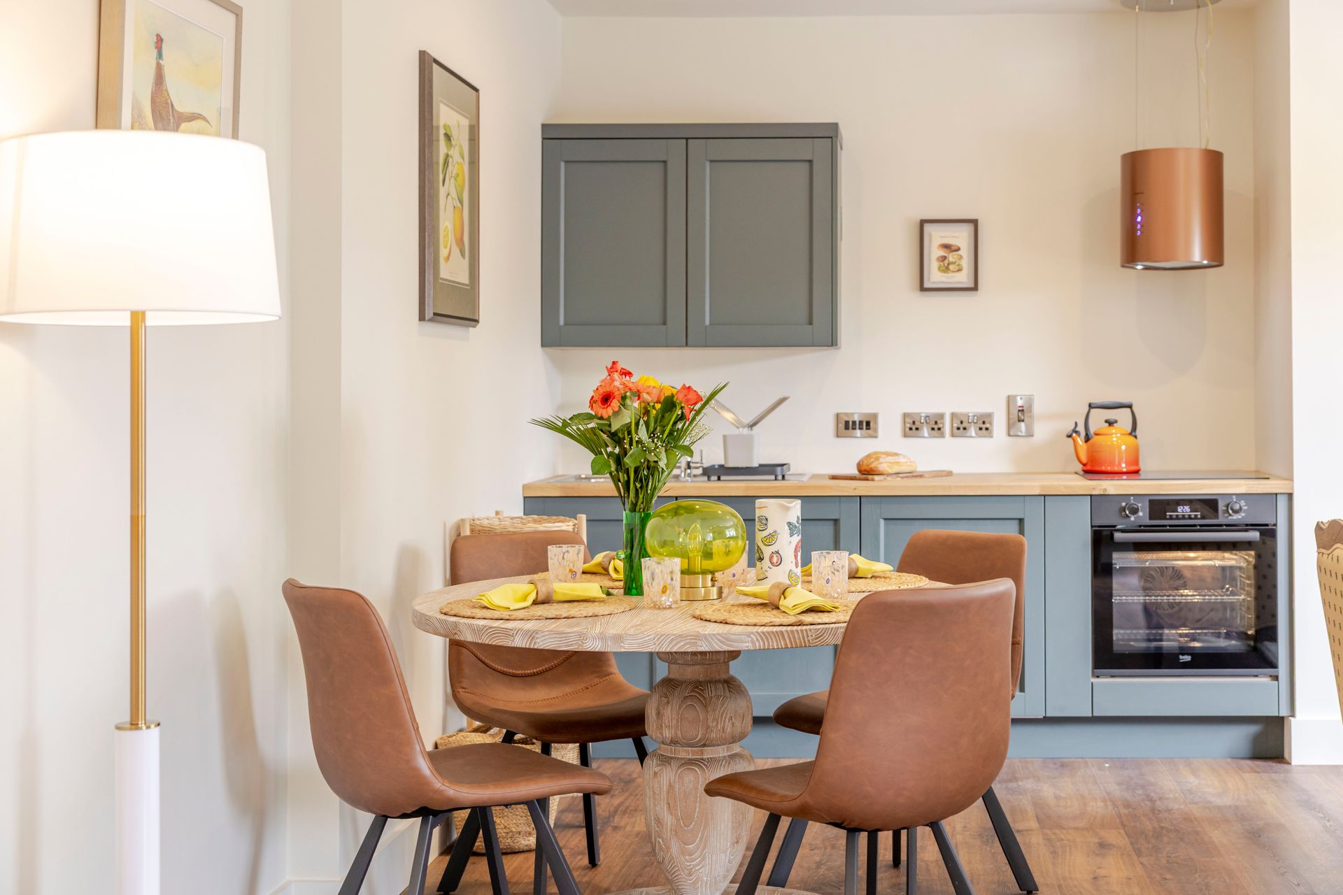 Dining area with round table, flowers, and chairs beside a small blue kitchen with cabinets and oven