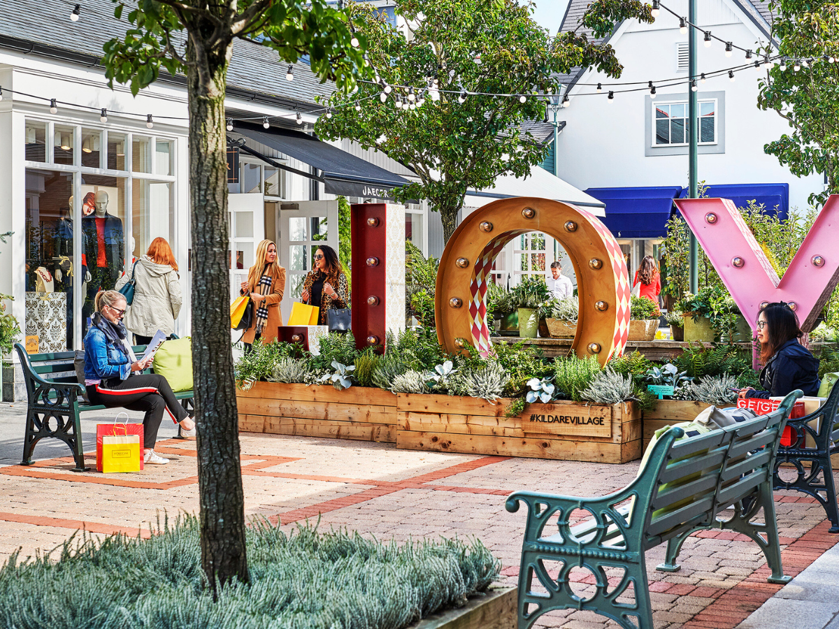 Outdoor plaza with large floral “JOY” letters, benches, trees, and people sitting nearby