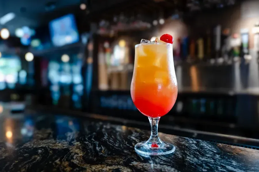A colorful layered tropical cocktail garnished with a cherry on a dark bar top.