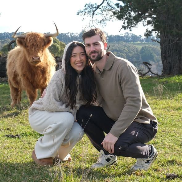 Two People Crouch Smiling in a Grassy Field With a Shaggy Brown Cow Behind Them — Lakeview Luxury Retreat in Canobolas, NSW