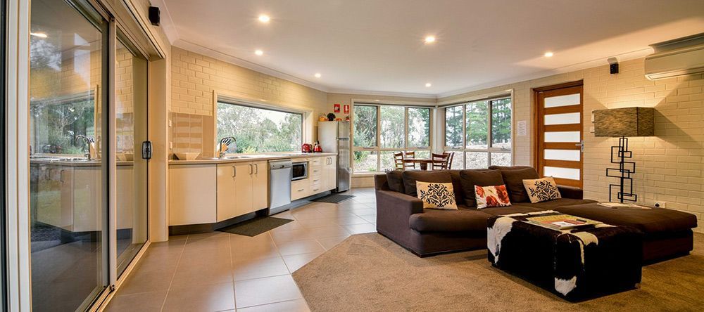 A Living Room With a Couch, Ottoman, and Sliding Glass Doors Leading to a Kitchen — Lakeview Luxury Retreat in Canobolas, NSW