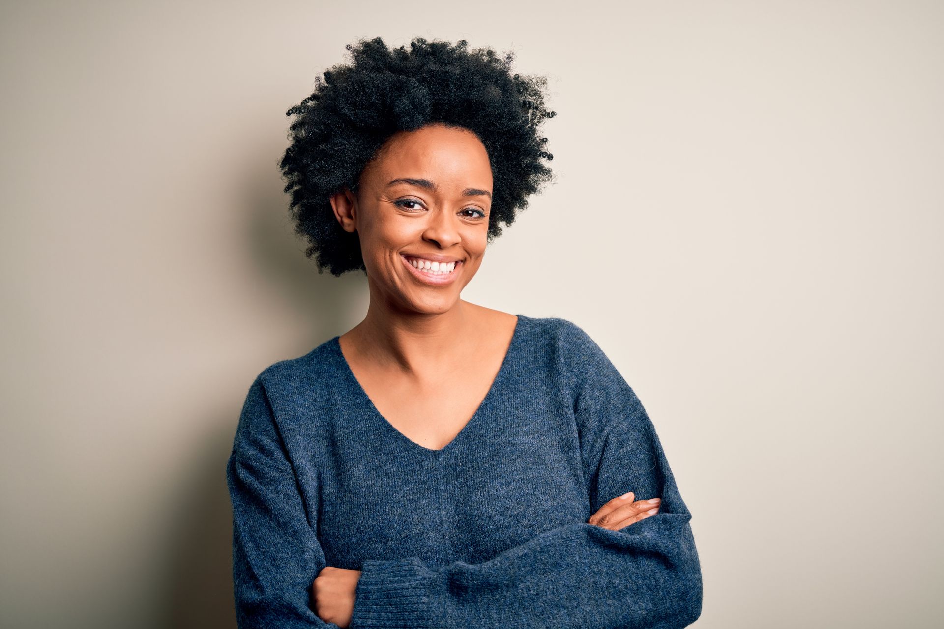 Smiling person with curly hair and crossed arms, wearing a dark blue sweater against a plain background