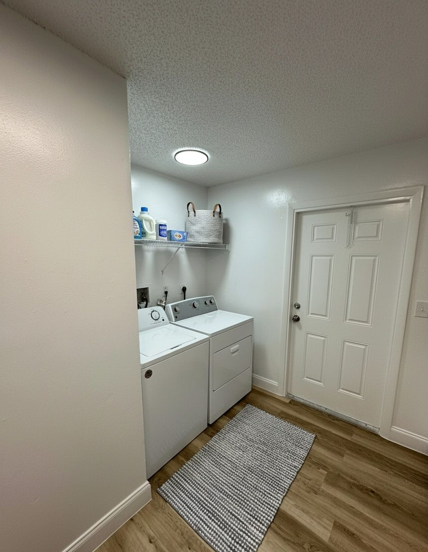 Laundry room with white appliances, shelving, and door. Gray rug on wood-look floor.