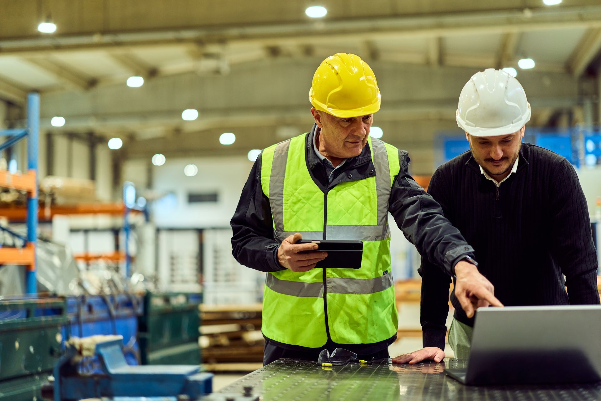 Two men in hard hats collaborate on a laptop while working in a warehouse environment.