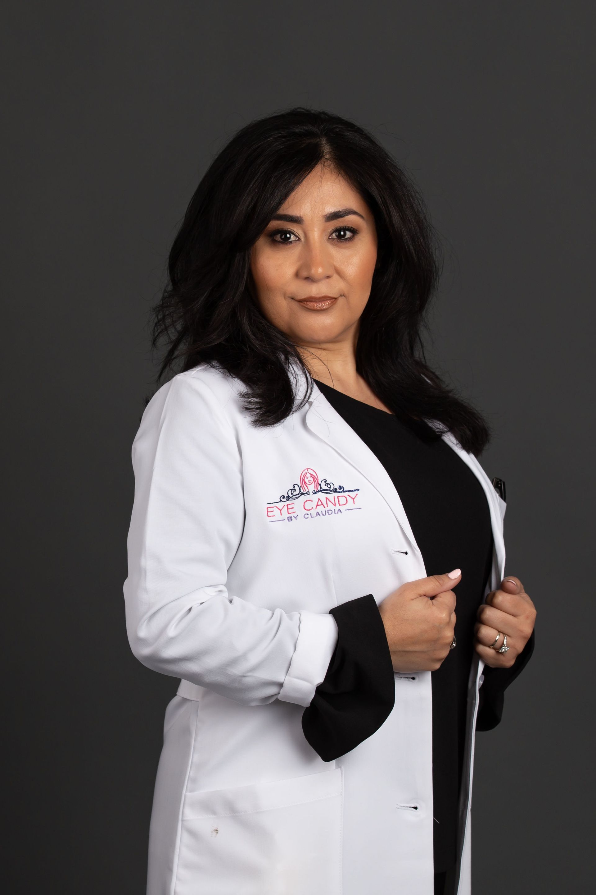 A woman in a white lab coat is standing in front of a gray background.