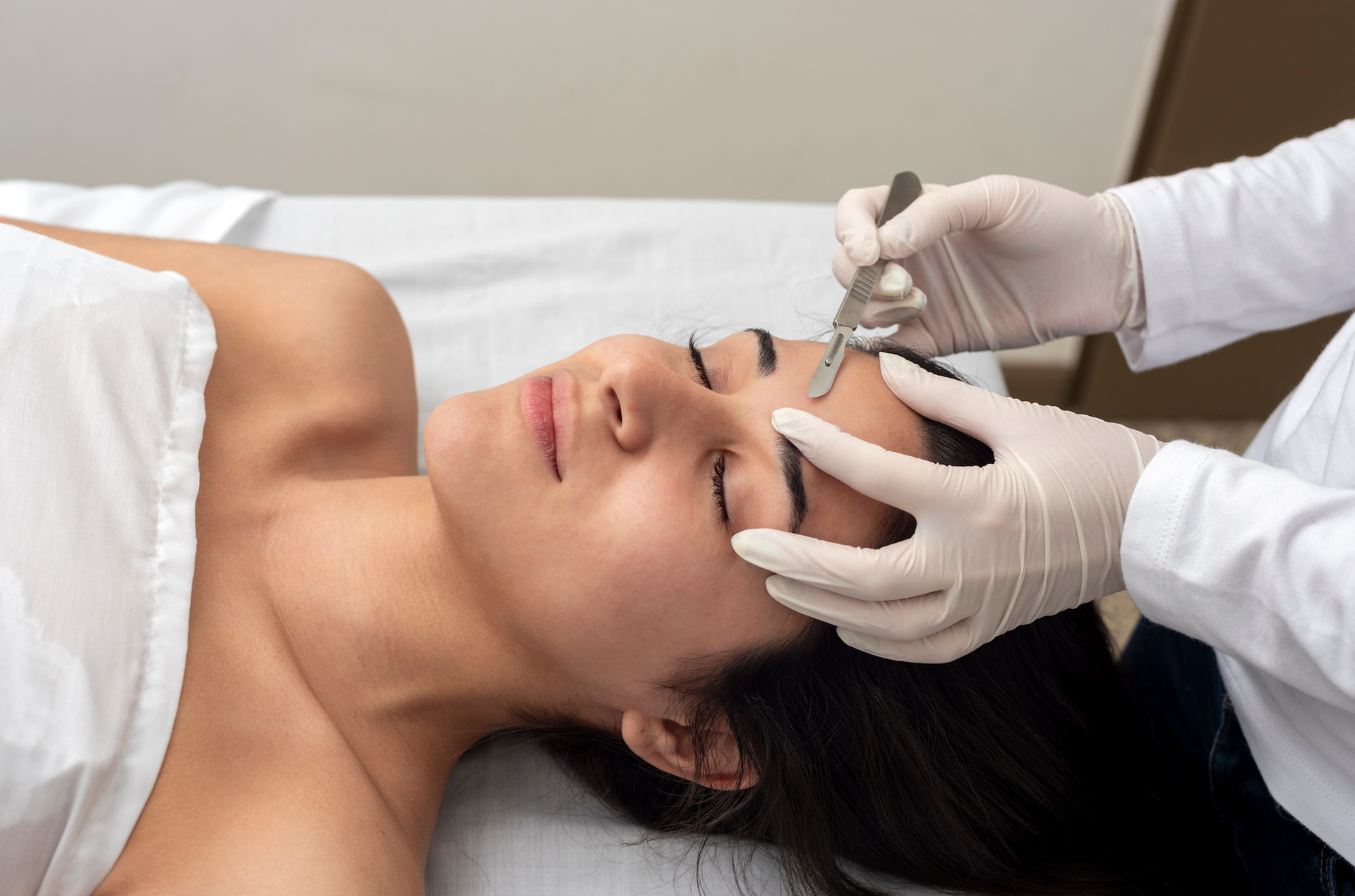 A woman is laying on a bed getting her eyebrows trimmed by a doctor.