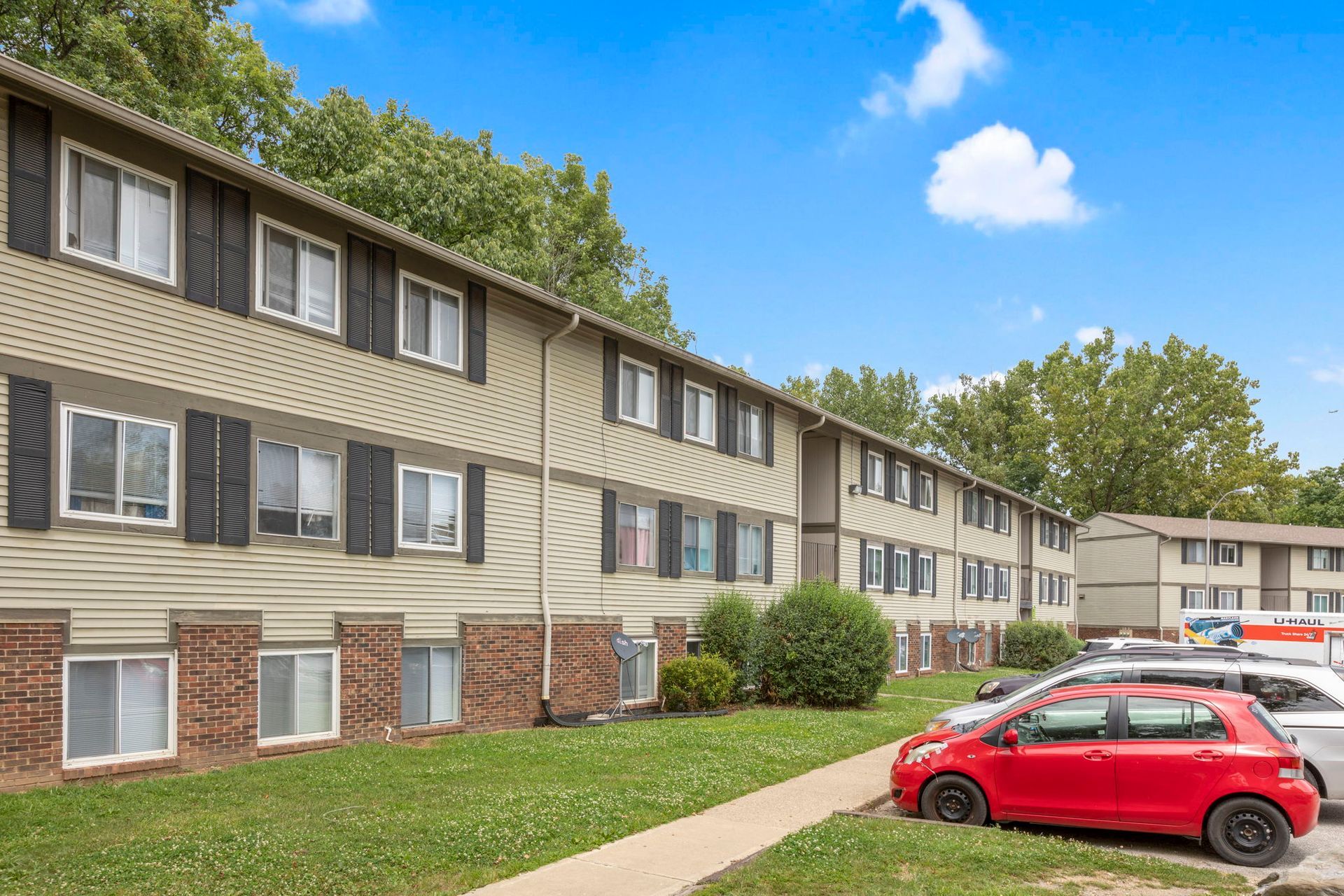 Apartment building with tan siding, red brick, and cars parked out front on a sunny day.